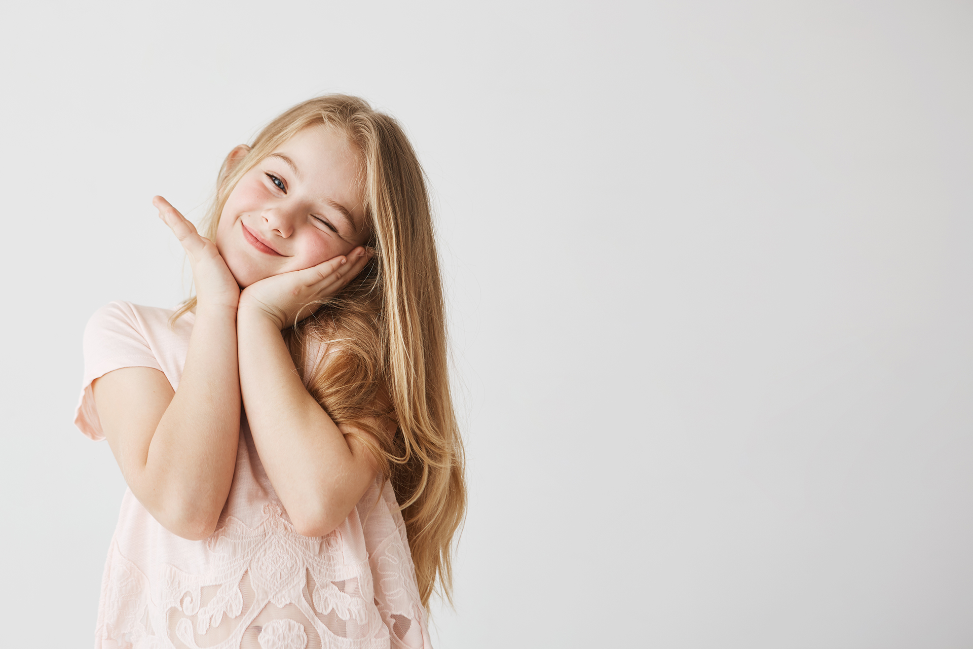 Beutiful little blonde girl smiles at camera winking, posing, touching face with her hands in pink cute dress. Child looking happy and delighted. Copy space